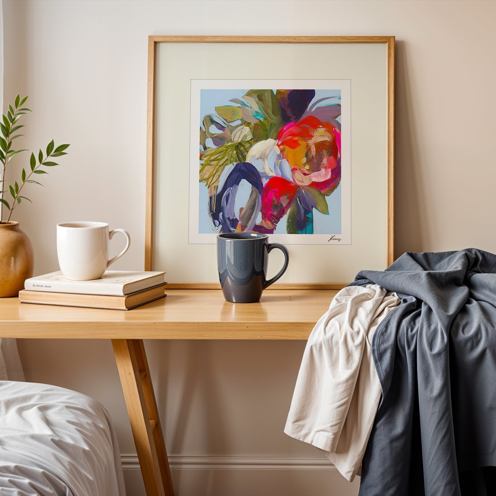 Framed abstract art on a wooden side table with books and mugs in a cozy room.