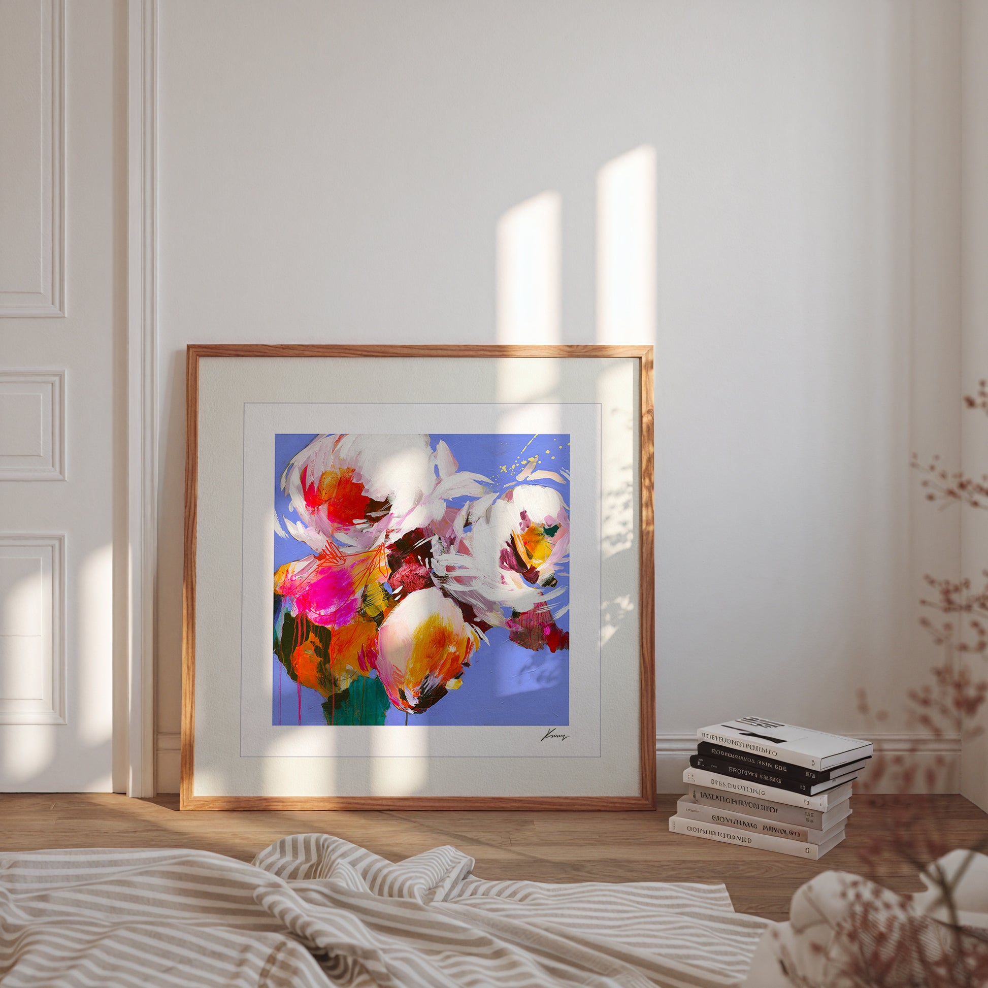 Framed abstract artwork on a wall with books and a blanket on a wooden floor.