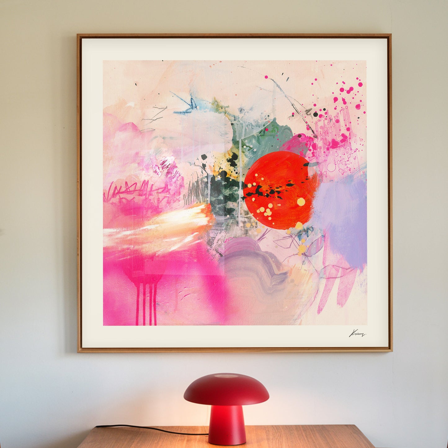 A vibrant abstract painting with pink and red tones, featuring a central red circle with white speckles, flanked by softer pink and blue strokes, presented in a gold frame, with a red lamp in the foreground.