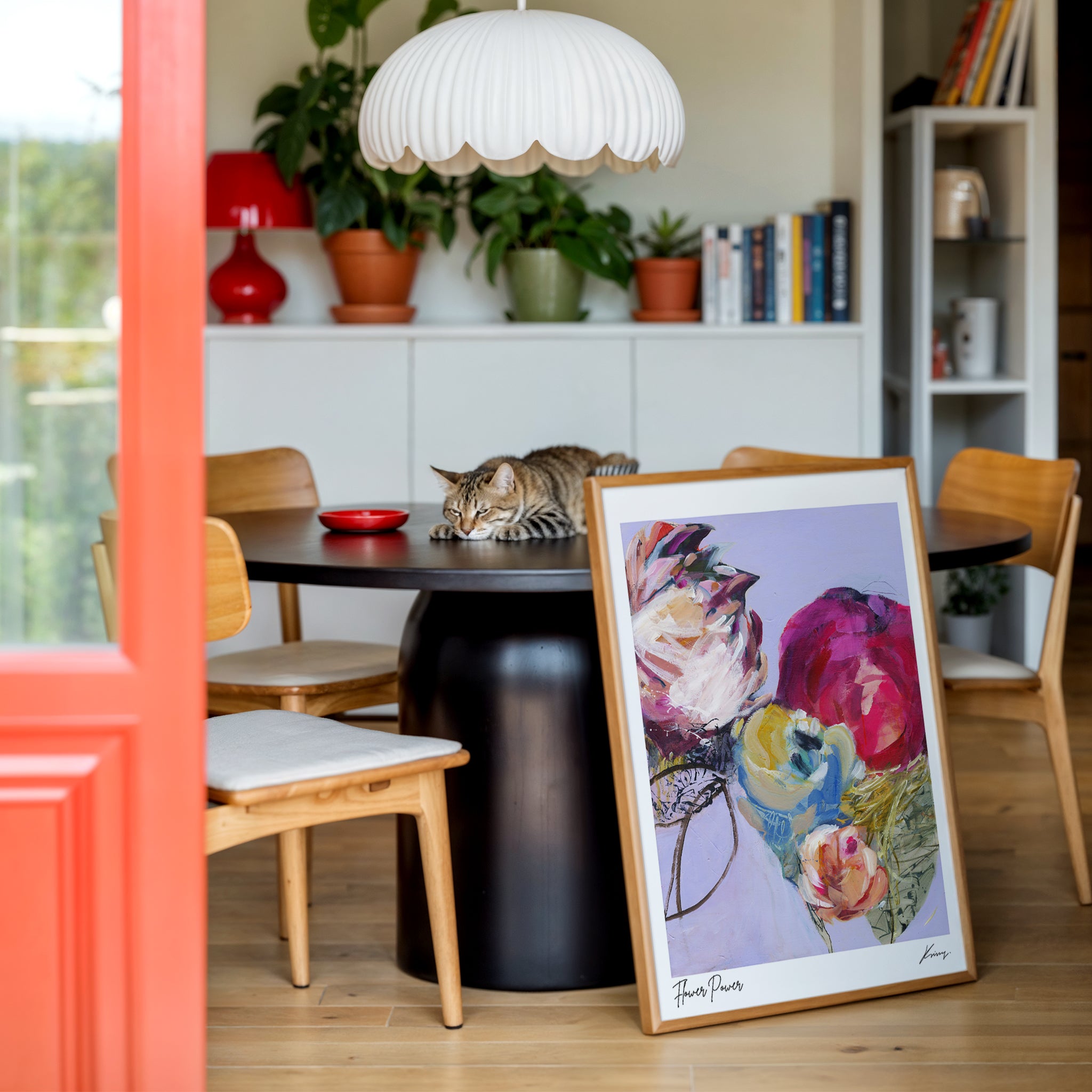 Dining room with a framed floral painting on a table, books, and potted plants.