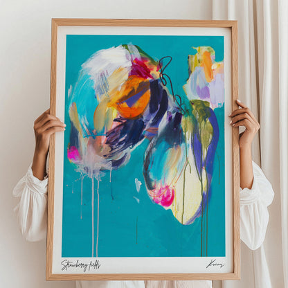 Person holding a colorful abstract painting in a wooden frame against a white wall.