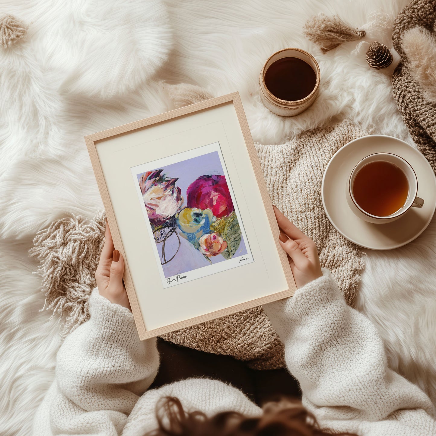 Person holding a framed floral print on a soft, white surface with a cup of tea.