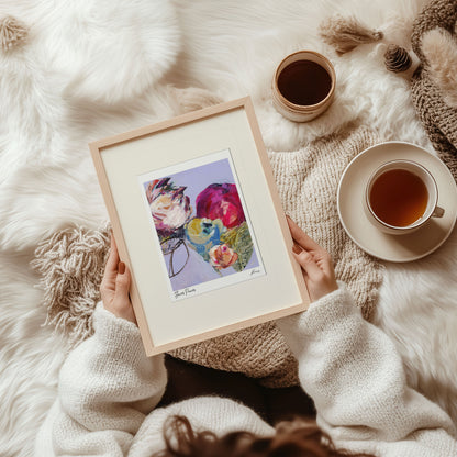 Person holding a framed floral print on a soft, white surface with a cup of tea.
