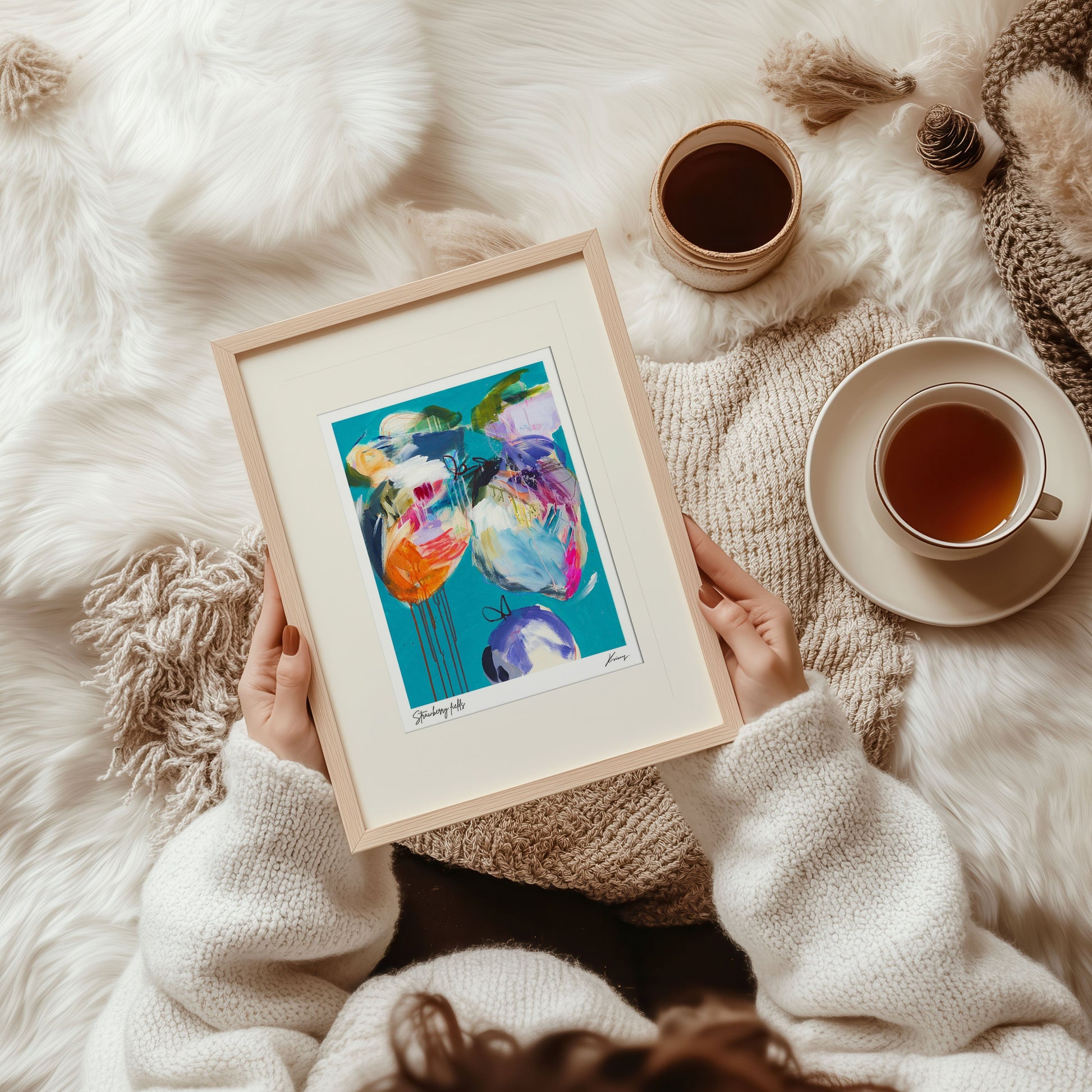 Person holding a framed colorful artwork on a soft, white surface with a cup of tea.