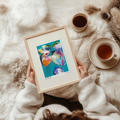 Person holding a framed colorful artwork on a soft, white surface with a cup of tea.