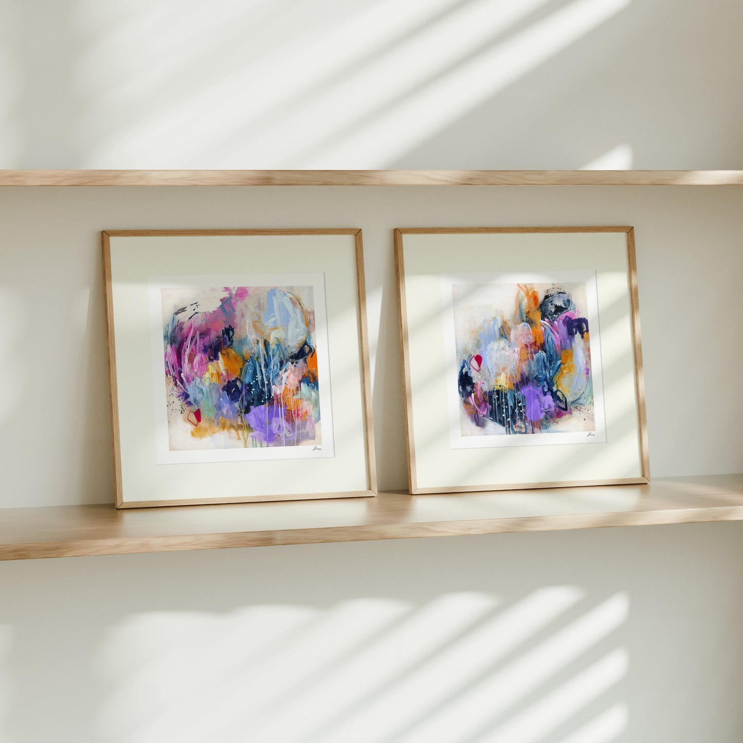 Two framed abstract artworks on a wooden shelf with sunlight casting shadows on a white wall.