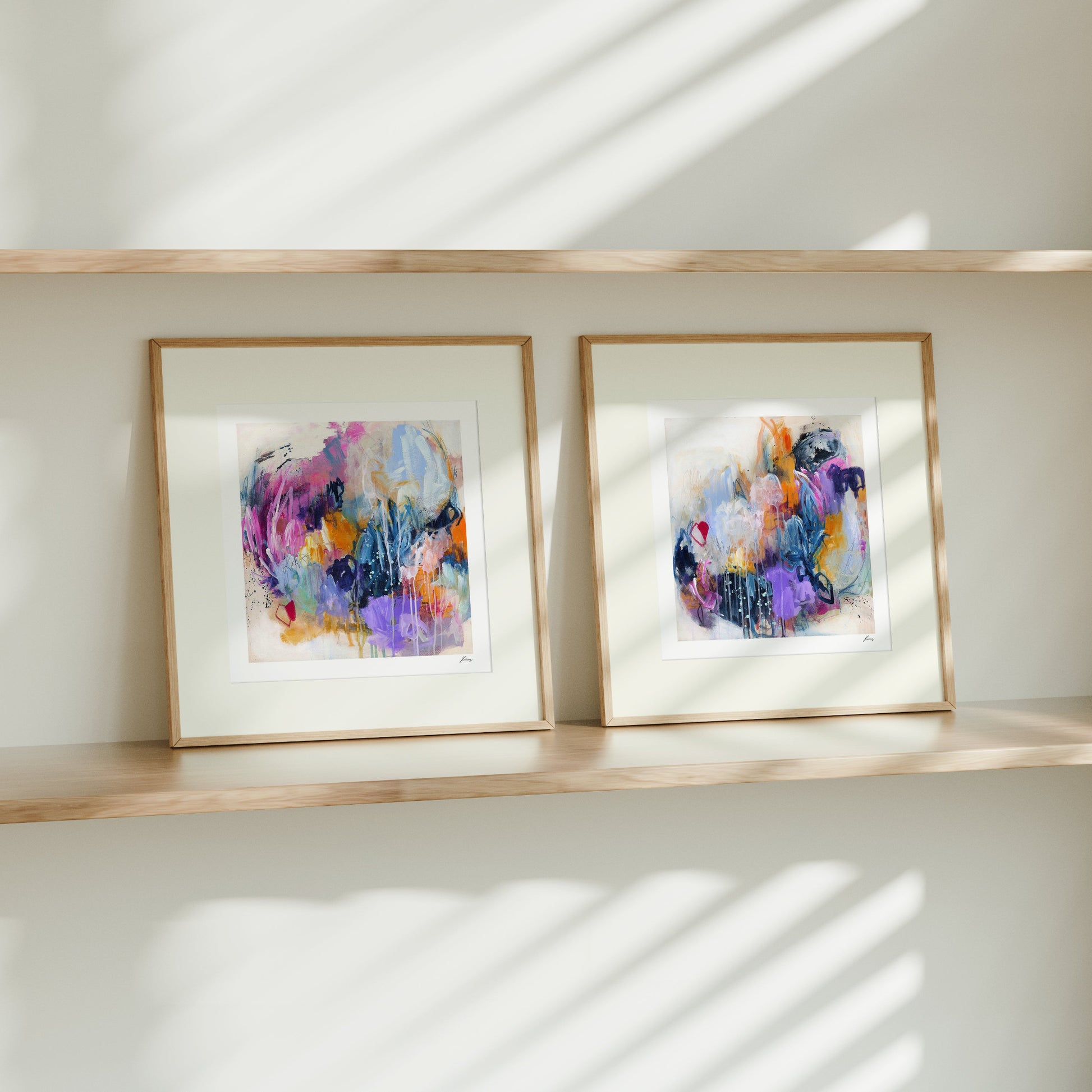Two framed abstract artworks on a wooden shelf with sunlight casting shadows on a white wall.