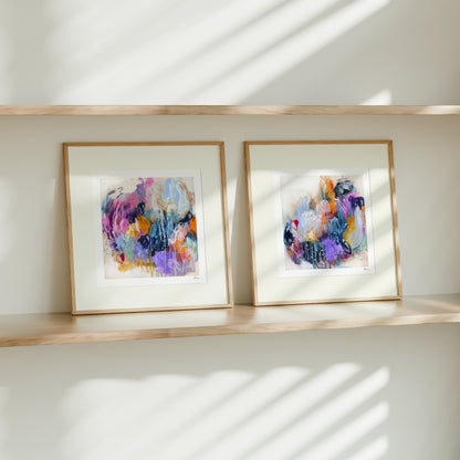 Two framed abstract artworks on a wooden shelf with sunlight casting shadows on a white wall.
