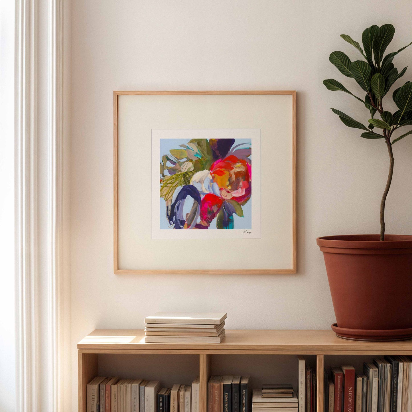 Framed abstract artwork on a wall above a bookshelf with a plant to the right.
