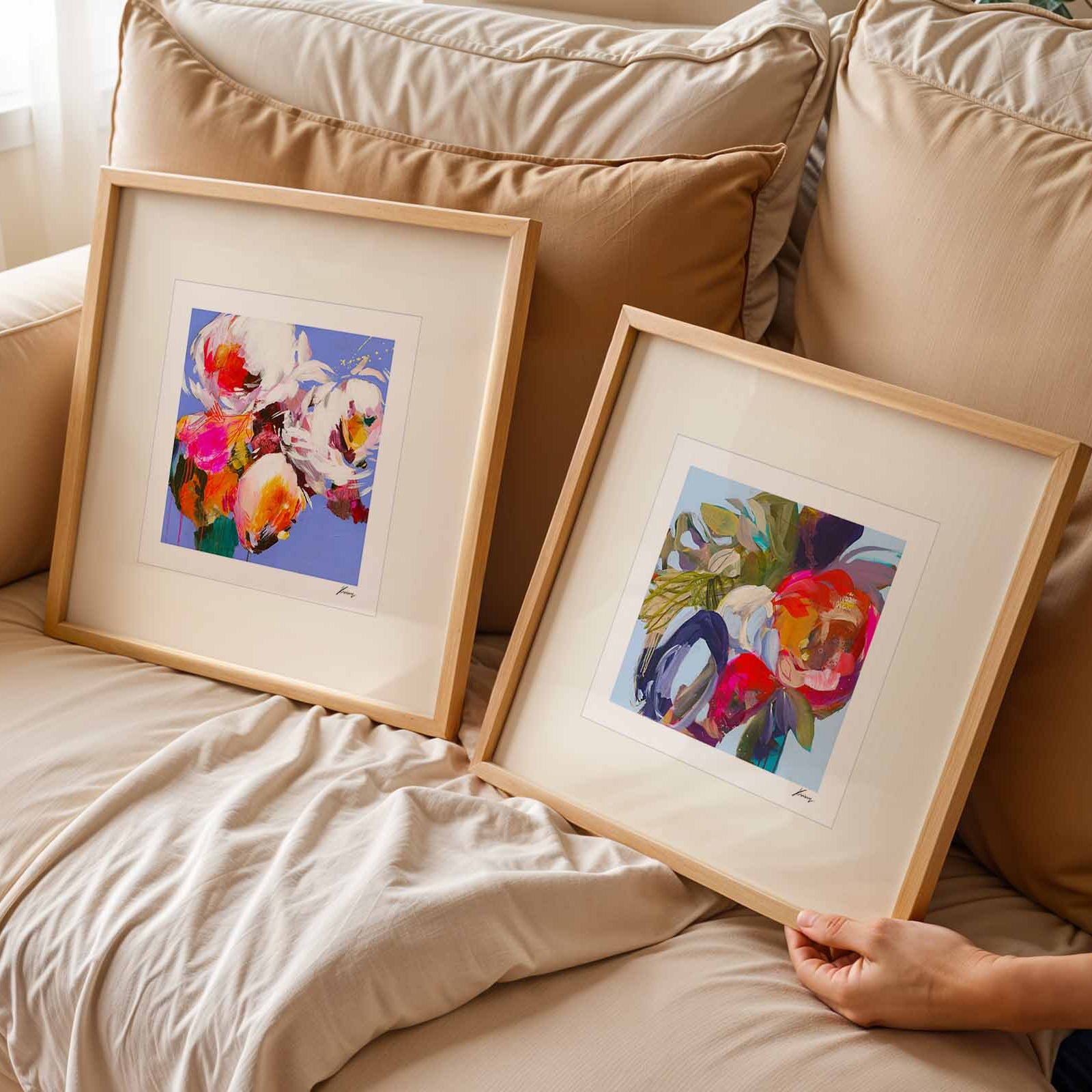Two framed artworks on a beige sofa with a person's hand reaching towards one of them.