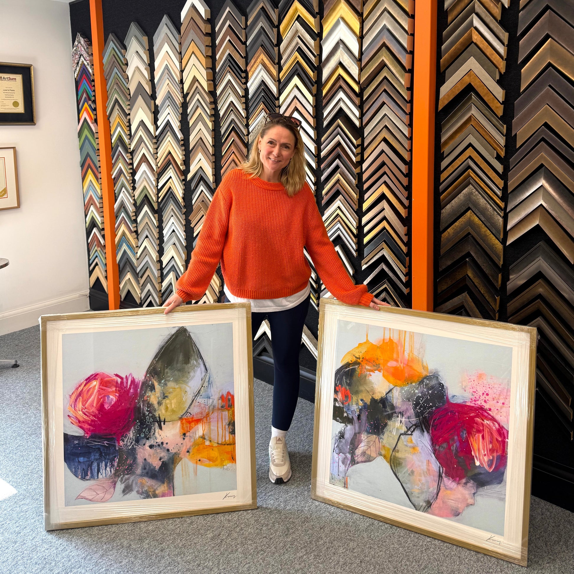 Woman holding two framed artworks in front of a wall of picture frames.