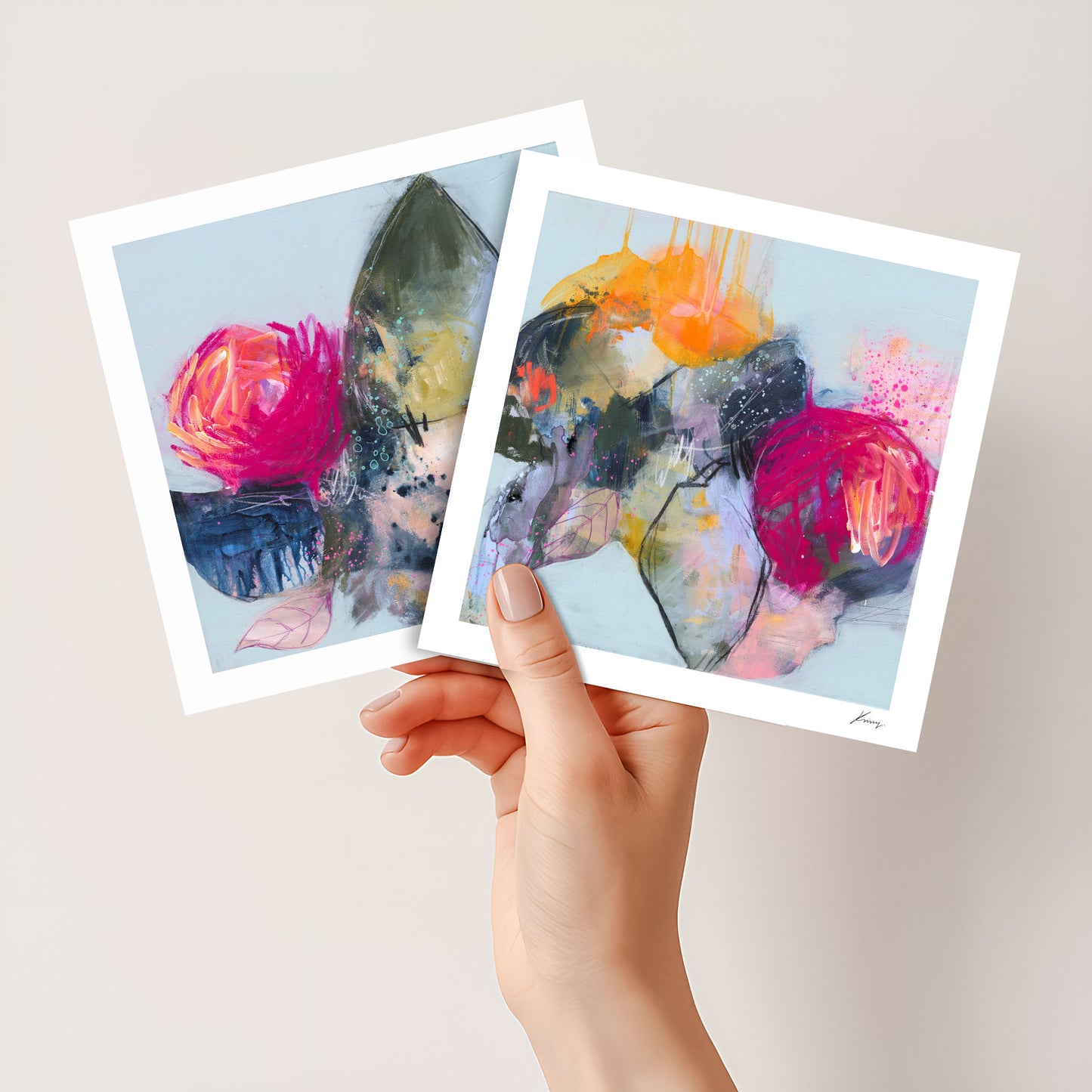 Hand holding two abstract art prints with colorful brush strokes on a light background